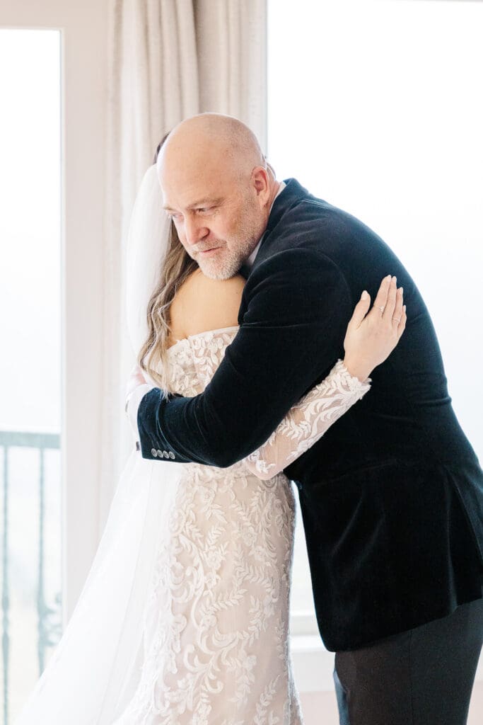 Father embraces bride in a heartfelt hug during a quiet moment by the window.
