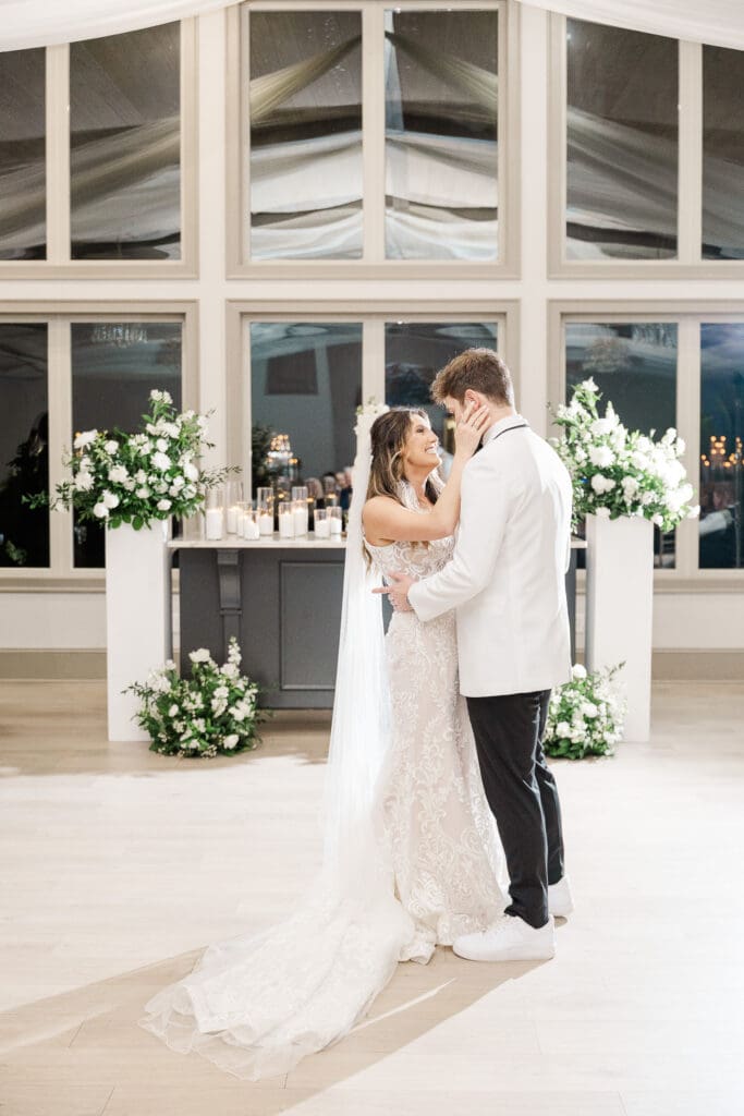 Bride and groom share a romantic first dance surrounded by candlelight and white floral arrangements.