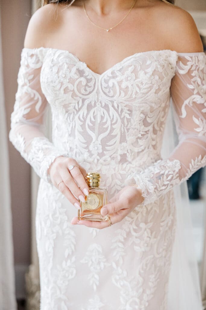 Bride in lace wedding gown holding gold perfume bottle during getting ready