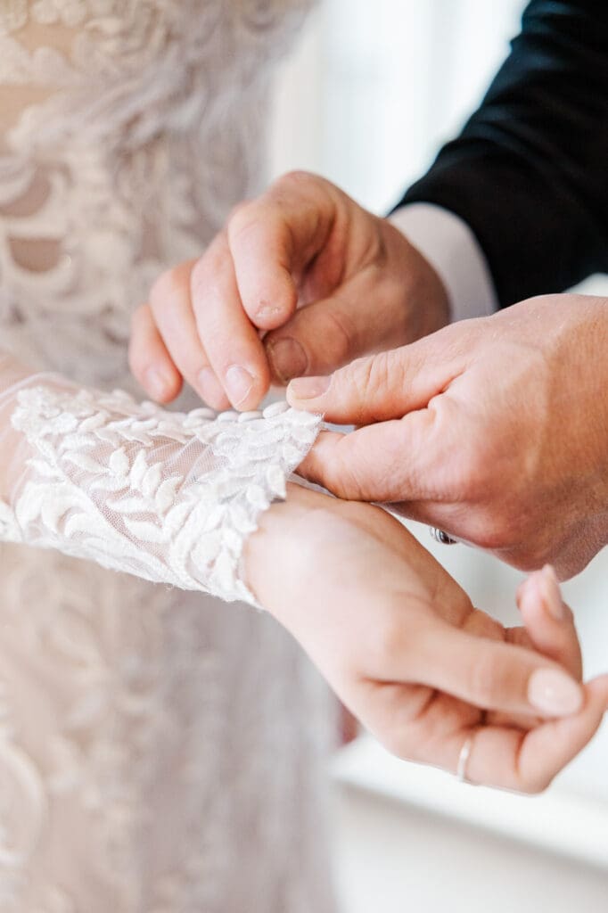 Close-up of father of the bride's hands fastening lace buttons on bride’s sleeve.