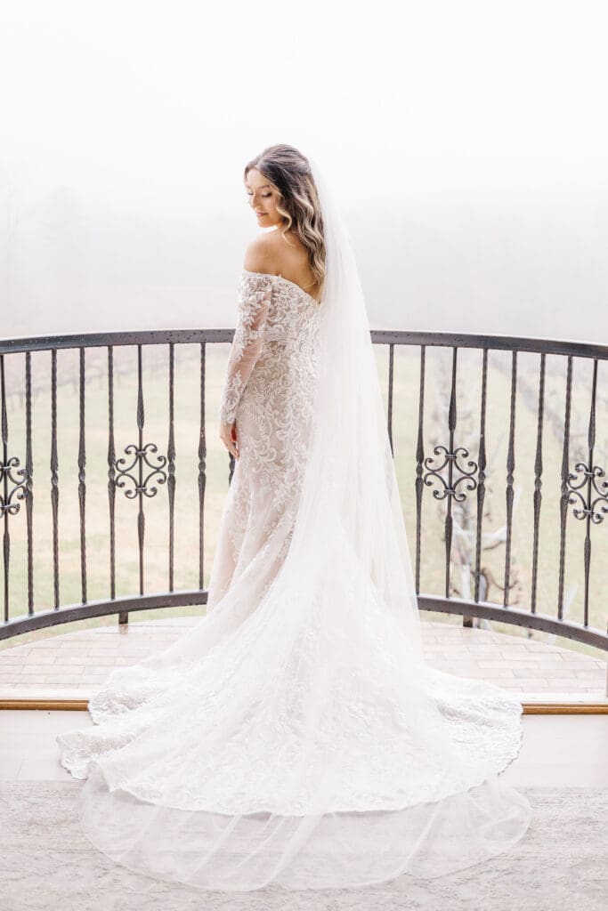 Bride standing on balcony in long lace gown with cathedral veil flowing at Venue In The Vines, Ellijay, GA