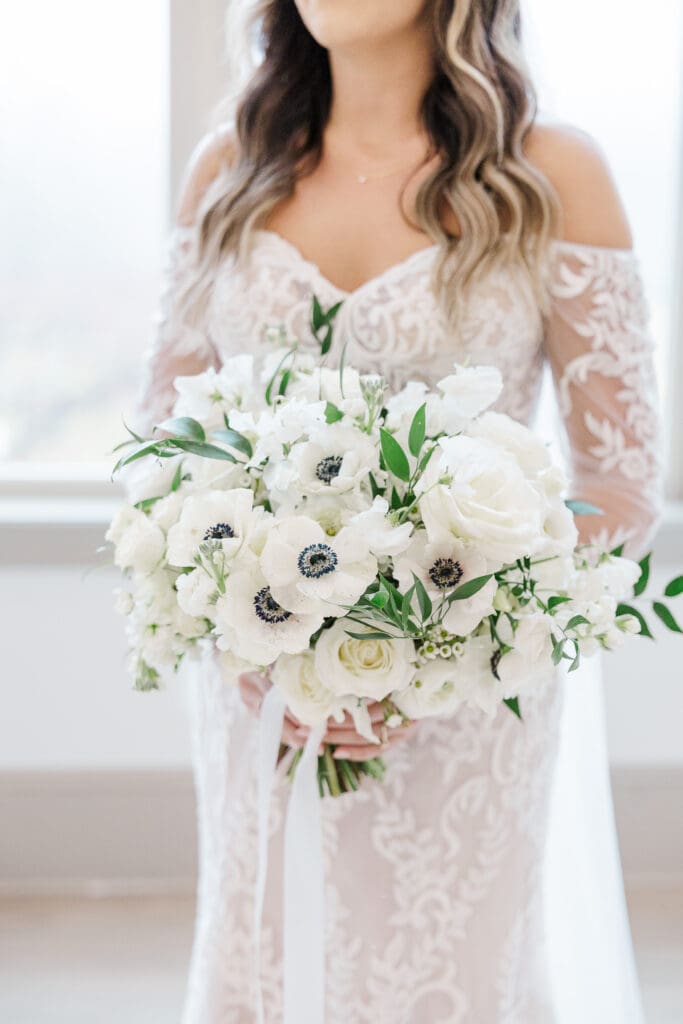 Bride holds a lush bouquet of white anemones, roses, and greenery in a lace off-shoulder gown. Bouquet designed my Marigo Flowers.