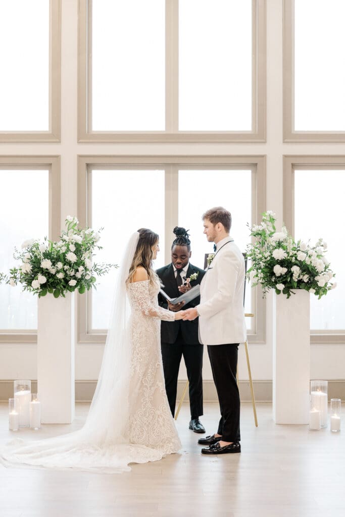 Bride and groom exchange vows in a light-filled indoor ceremony framed by tall windows, glowing candles, and lush floral arrangements.