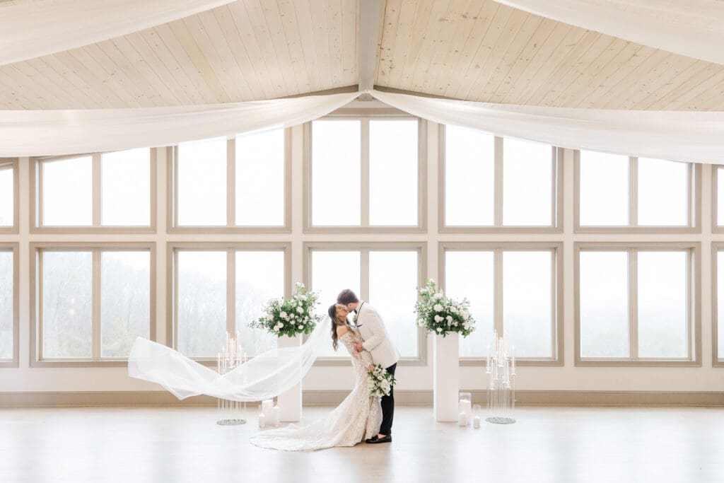 Bride and groom kiss in front of tall windows at Venue In The Vines in Ellijay, GA, with white florals and candles. Photo by Shanle Bowen, an Atlanta Wedding Photographer.