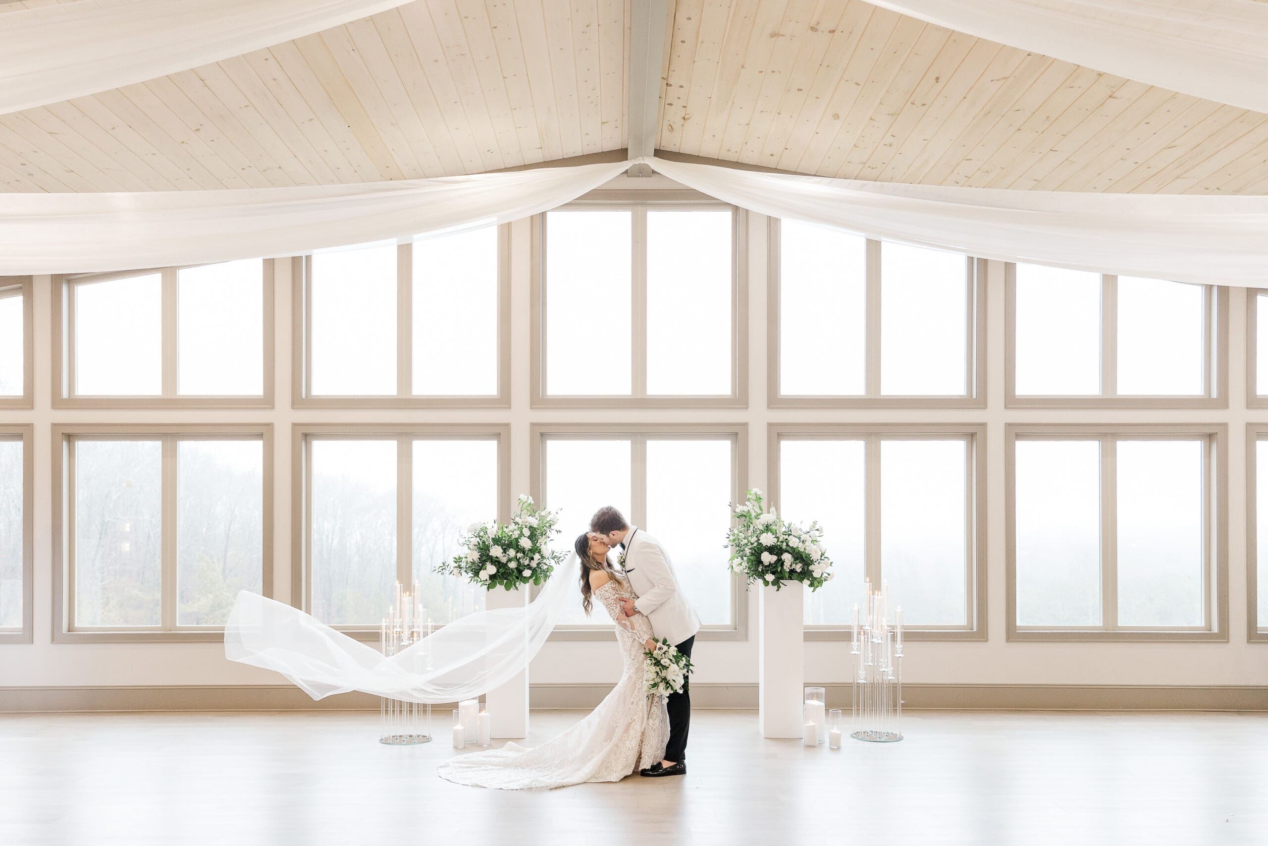 Bride and groom kiss in front of tall windows at Venue In The Vines in Ellijay, GA, with white florals and candles. Photo by Shanle Bowen, an Atlanta Wedding Photographer.