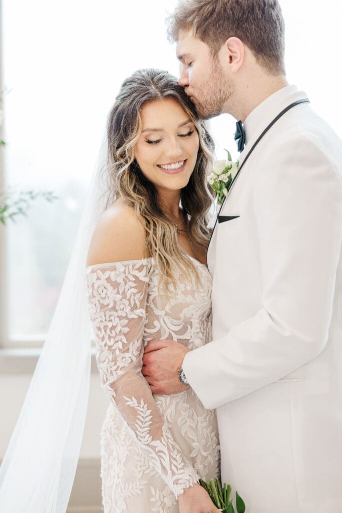 Groom kisses bride’s forehead as she smiles softly. Bride is wearing a beautifully detailed lace gown.