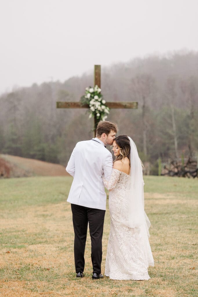 Bride and groom share a quiet moment under a large wooden cross with white floral accents on the vineyard lawn.