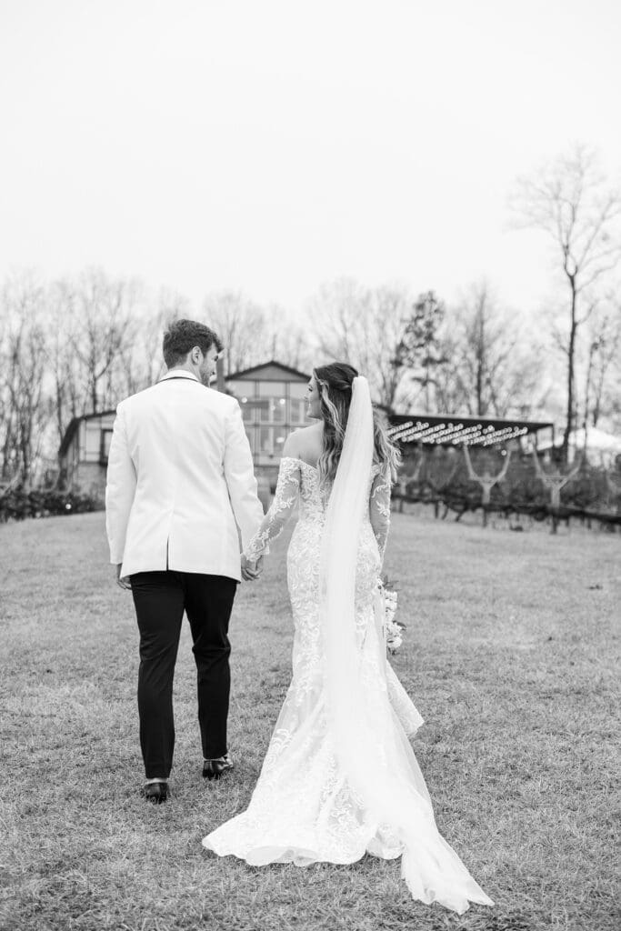Bride and groom walk hand in hand across vineyard lawn with twinkle lights in background at Venue In The Vines.