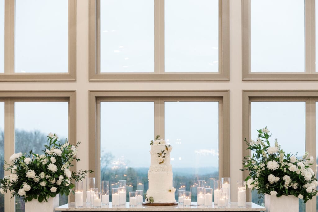 Elegant white wedding cake surrounded by candles and floral arrangements in front of large windows.