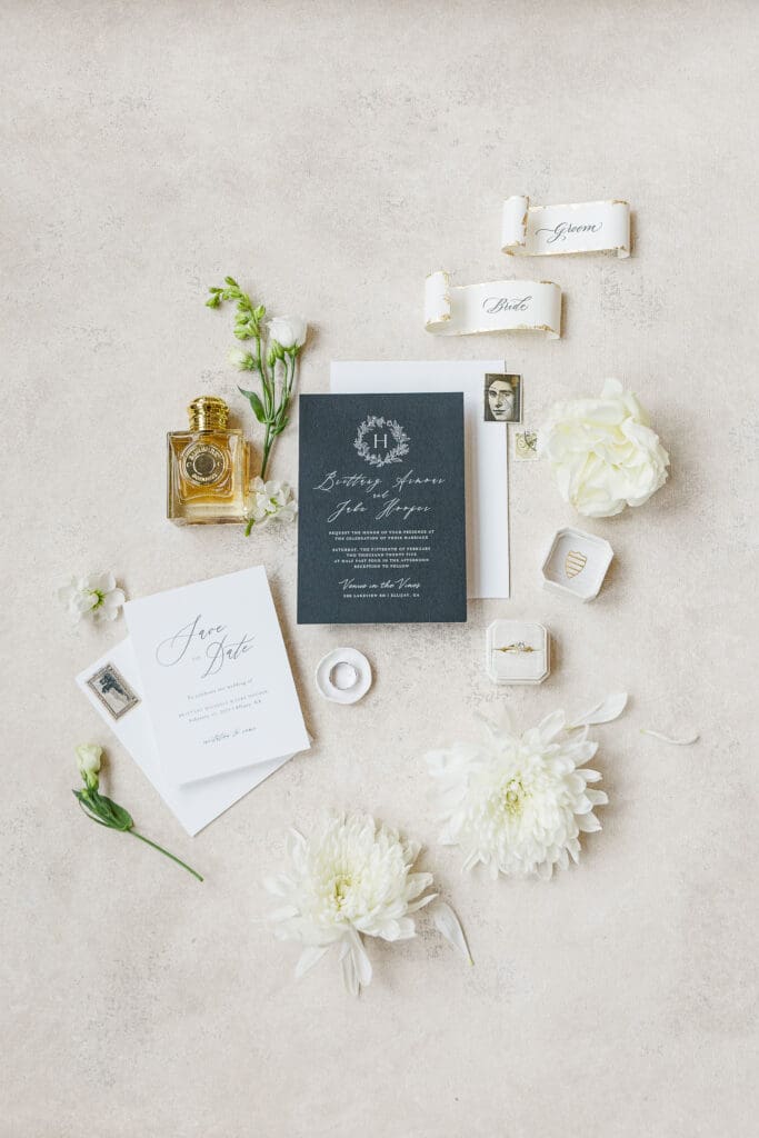 flat lay of white-on-black invitations, perfume, and white flowers on textured canvas backdrop by Chasing Stone.