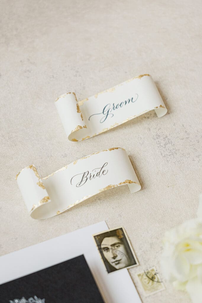 Bride and groom name cards with gold leaf detail styled flat lay on neutral linen surface.