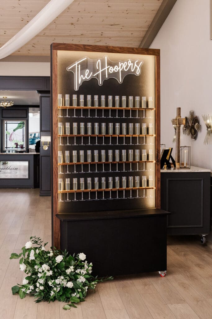 Champagne wall with neon ‘The Hoopers’ sign and glasses at wedding reception at Venue In The Vines.