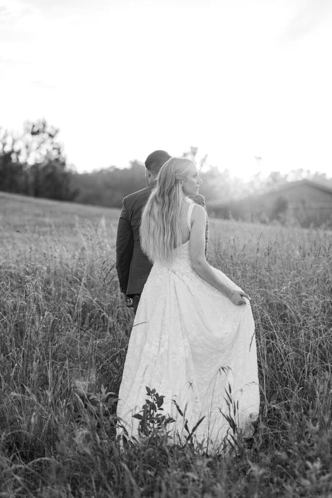 Bride and groom walking hand in hand through a sunlit field at sunset, bride holding her dress. Photo by Shanle Bowen, Atlanta GA Wedding & Engagement Photographer.