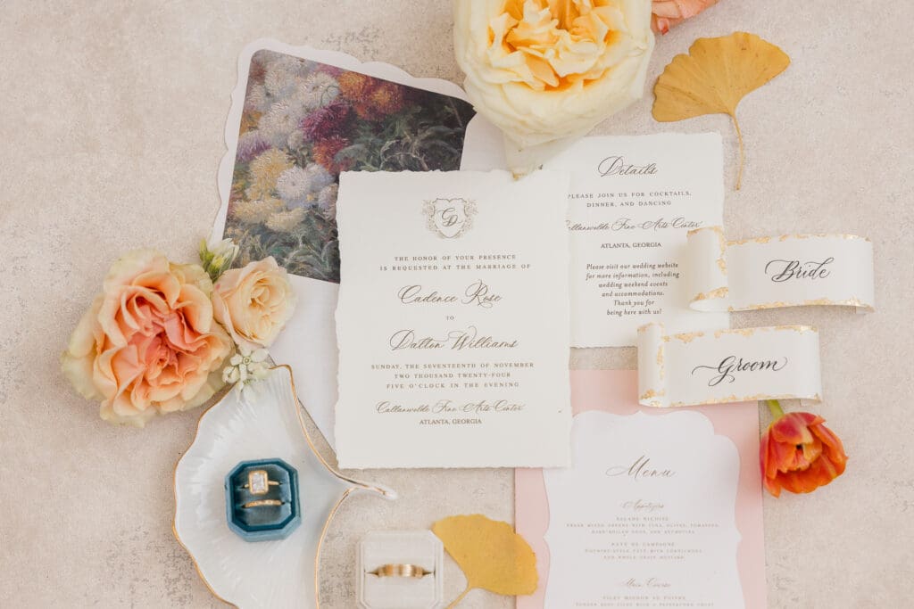 Flat lay of elegant wedding invitations, garden roses, and rings styled with peach and cream tones. 